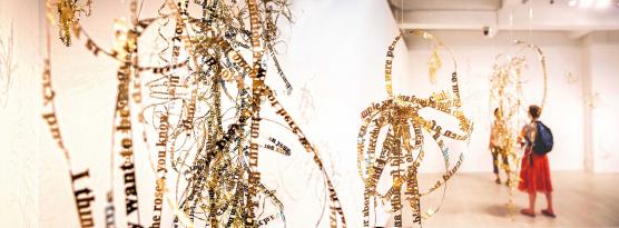 gold letters make up sentences that are tied in knots and hung from the ceiling of a gallery space