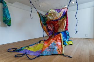Large unruly looking multicoloured painted sculptural form, sitting on the floor with it’s top half being hoisted into the air and suspended from ceiling with rope like tendrils