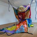 Large unruly looking multicoloured painted sculptural form, sitting on the floor with it’s top half being hoisted into the air and suspended from ceiling with rope like tendrils