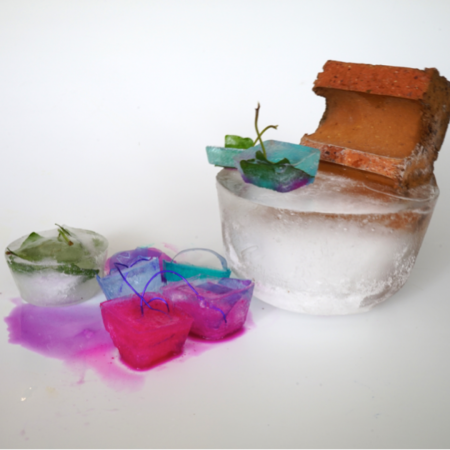 Some pink and purple coloured ice cubes next to a clear coloured ice cube topped with a brick